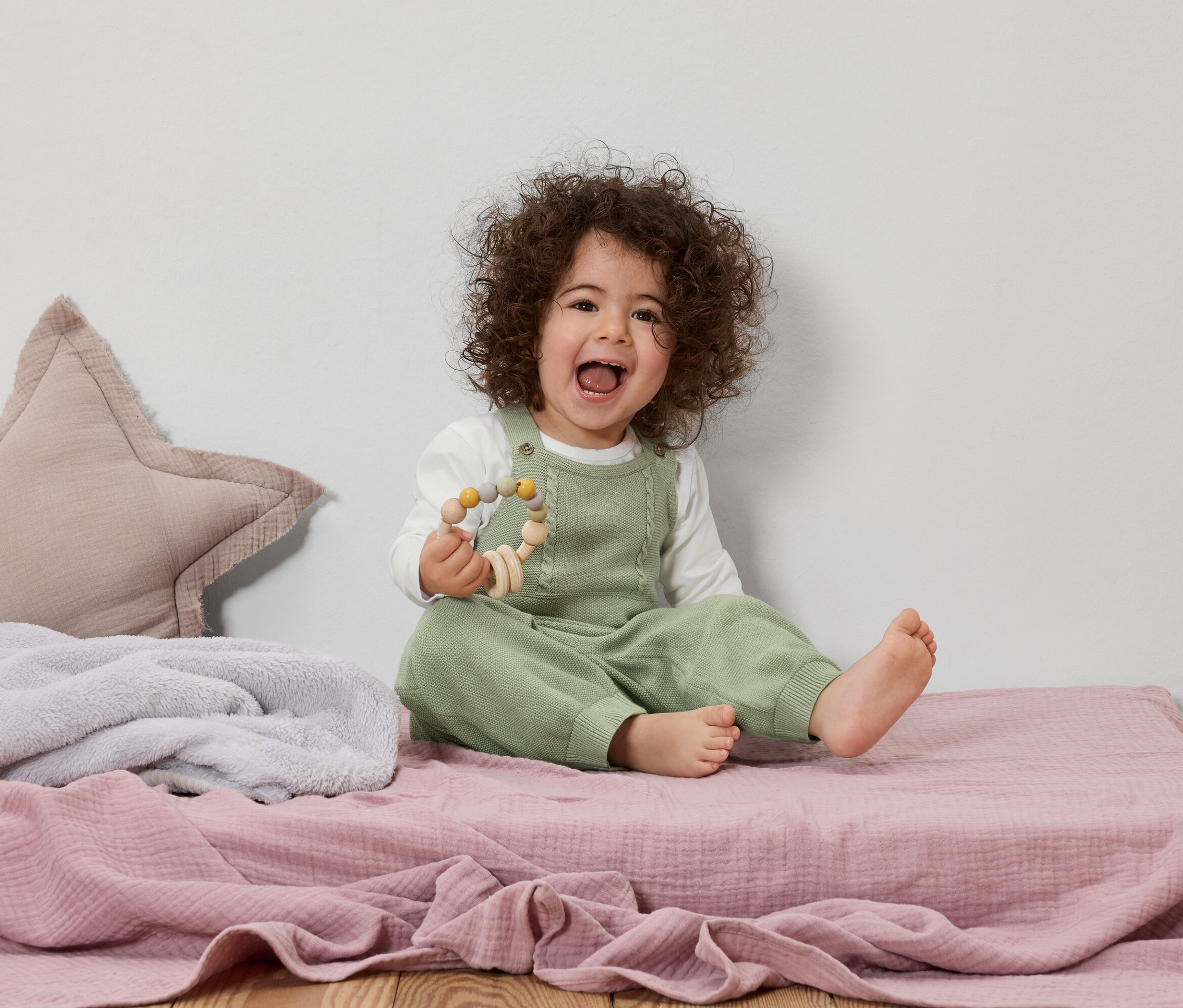 Uśmiechnięte dziecko z kręconymi włosami siedzi na różowym kocyku, trzymając drewnianą zabawkę.