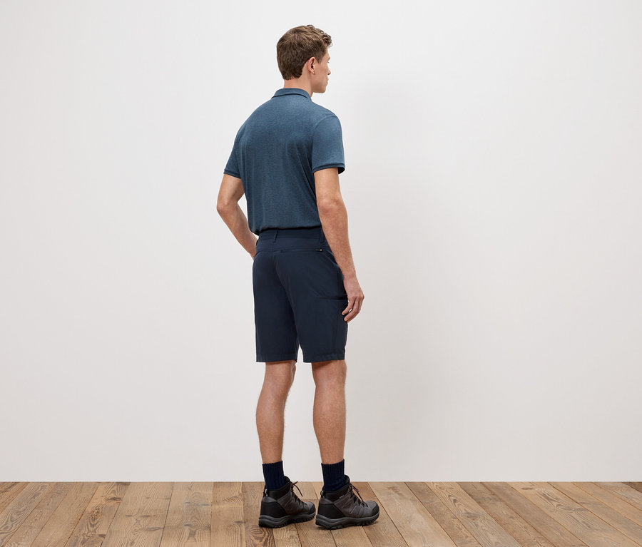 Mężczyzna stoi tyłem do kamery, ma na sobie niebieską koszulkę polo, szorty i buty trekkingowe na tle białej ściany.