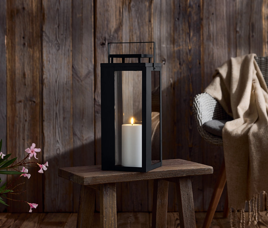 Czarny lampion ze świecą na drewnianym stole. W tle drewniana ściana i krzesło z kocem.