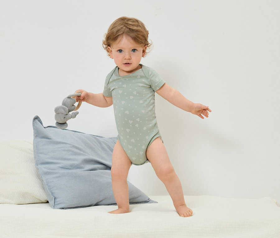 Małe dziecko w zielonym body w serduszka stoi na łóżku i trzyma szarą zabawkę słonia.