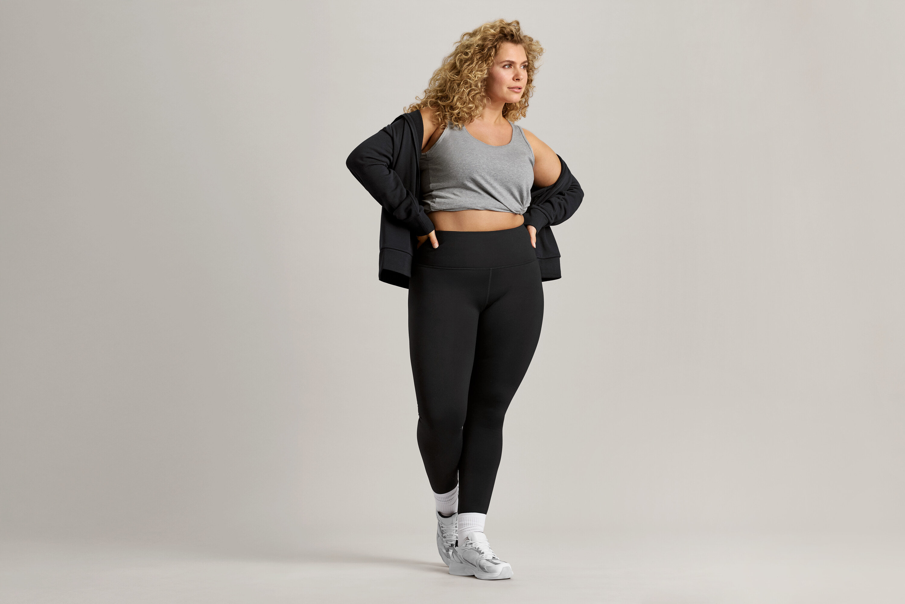 Kobieta o kręconych włosach ma na sobie szary tank top, czarną kurtkę i czarne legginsy.