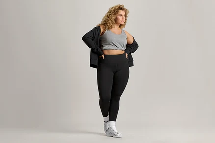 Kobieta o kręconych włosach ma na sobie szary tank top, czarną kurtkę i czarne legginsy.