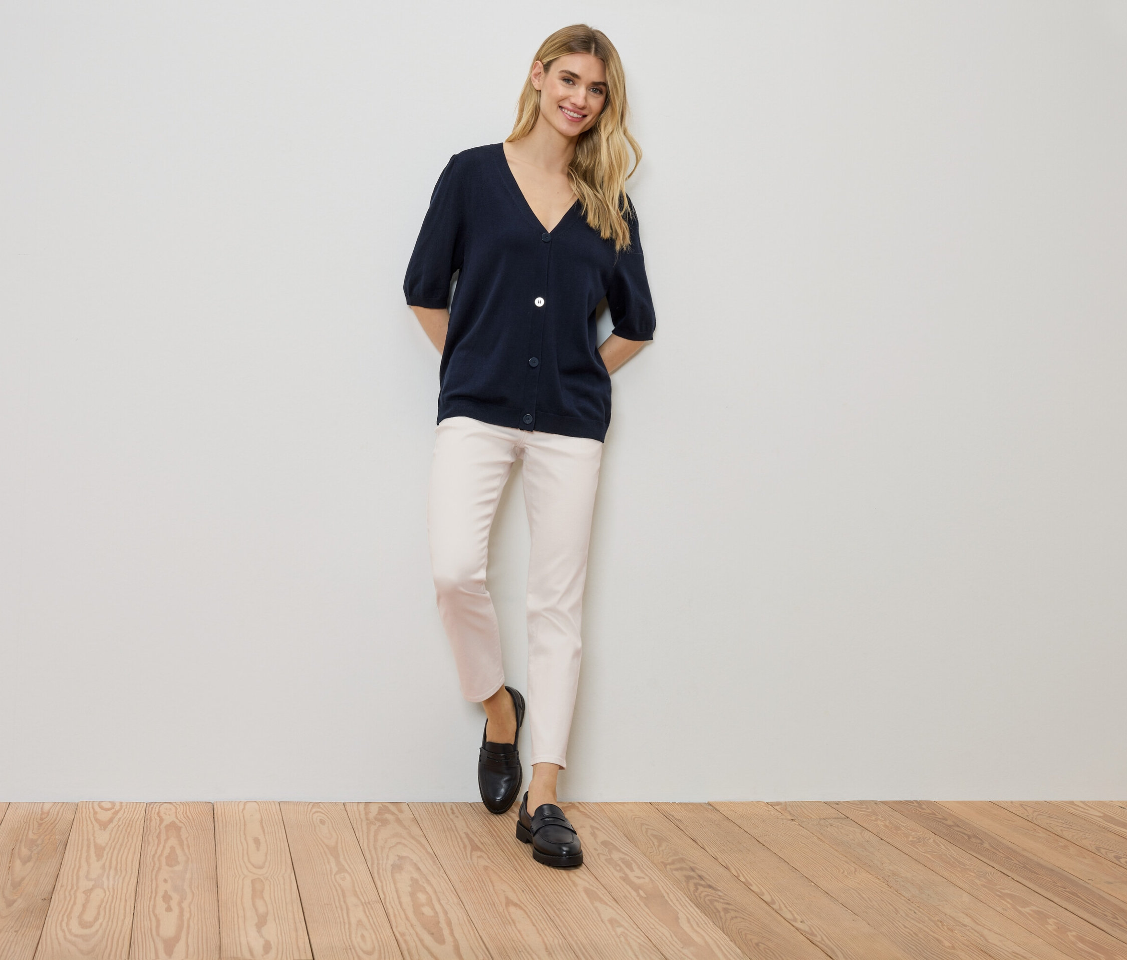 Kobieta stoi przy ścianie, ubrana w granatowy kardigan, jasne spodnie i czarne buty.