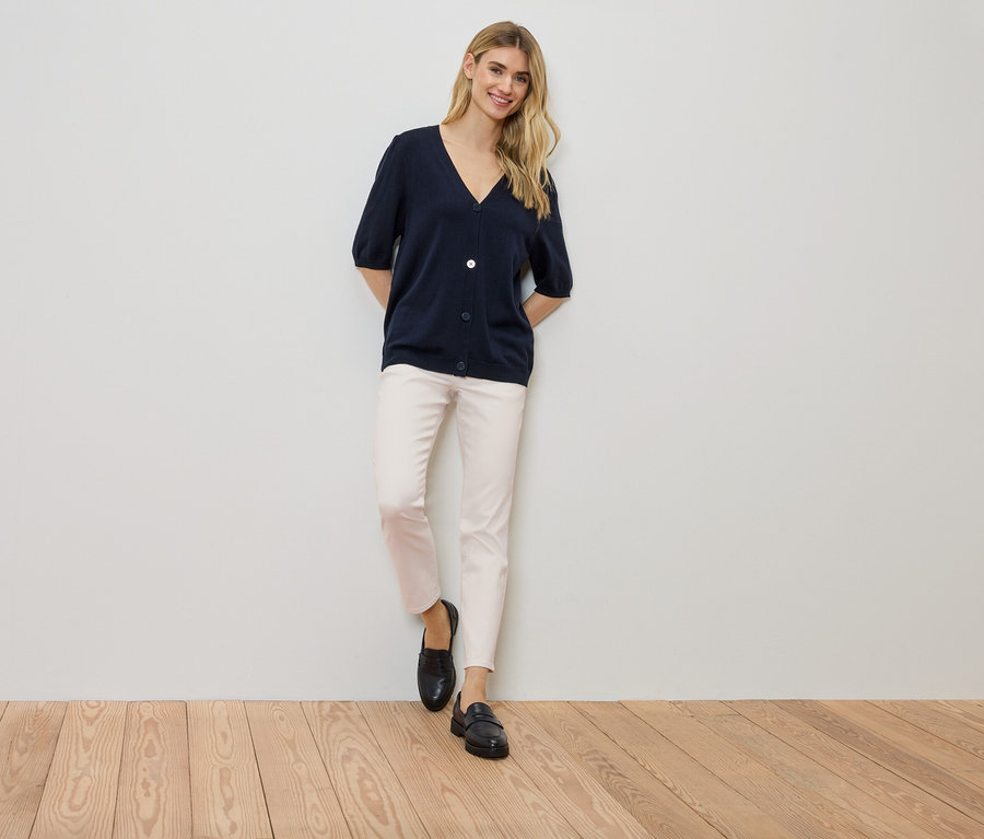 Kobieta stoi przy ścianie, ubrana w granatowy kardigan, jasne spodnie i czarne buty.