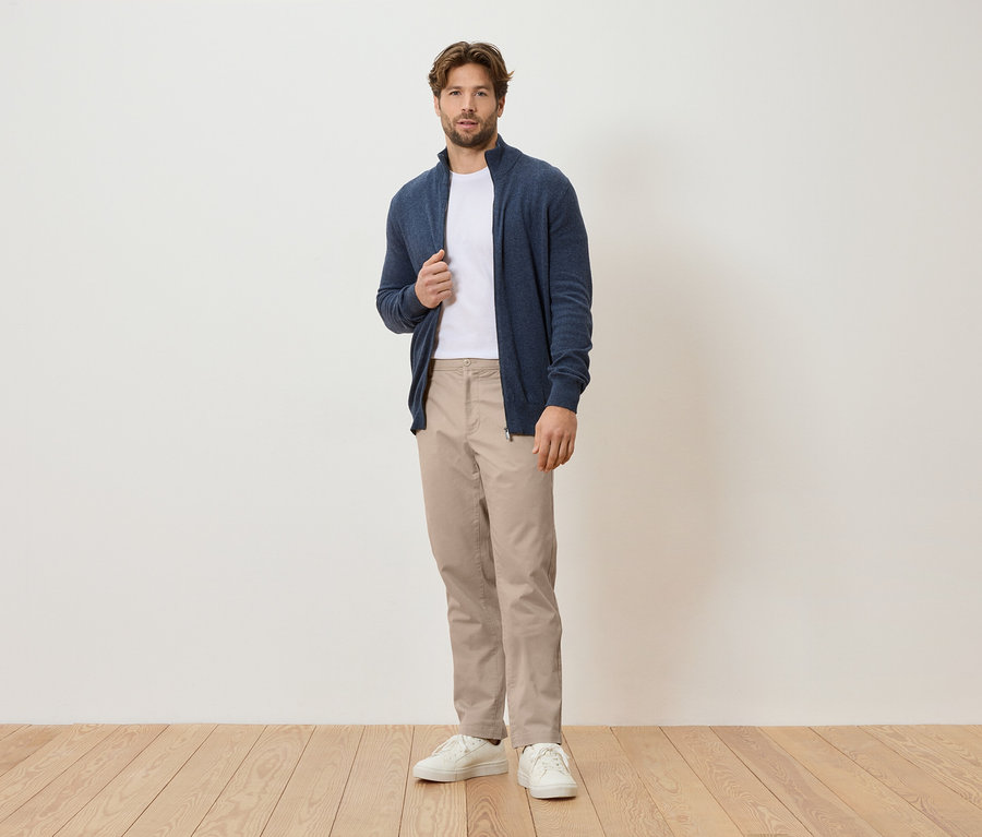 Mężczyzna stoi na tle białej ściany, ubrany w granatowy kardigan, biały t-shirt, beżowe spodnie i białe tenisówki.