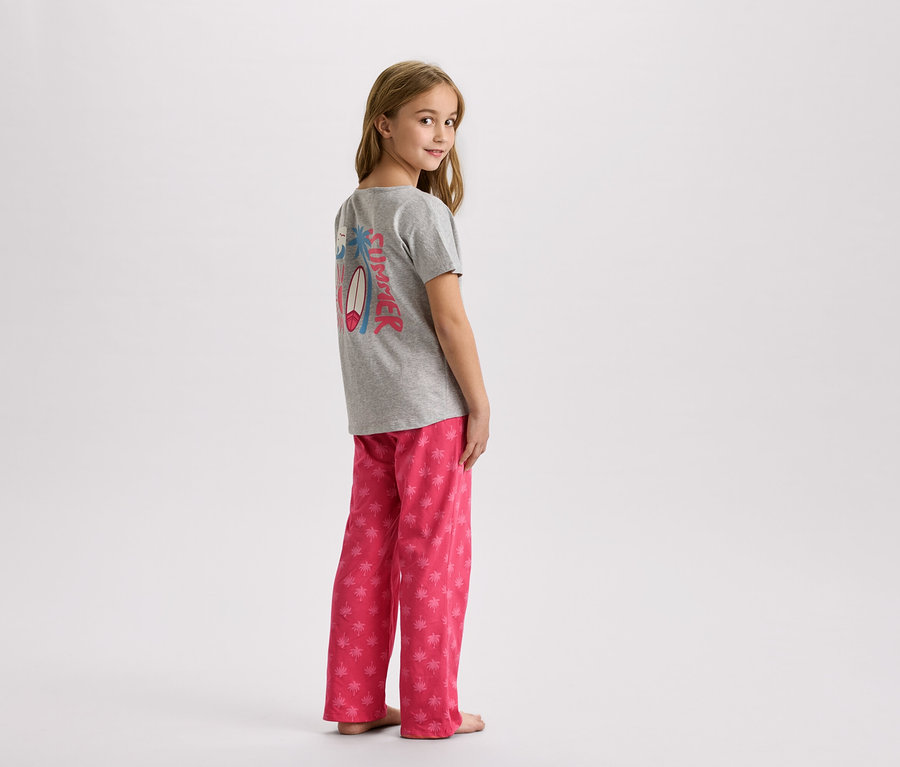 Młoda dziewczyna stoi w szarej koszulce z letnim wzorem i różowych spodniach w palmy.