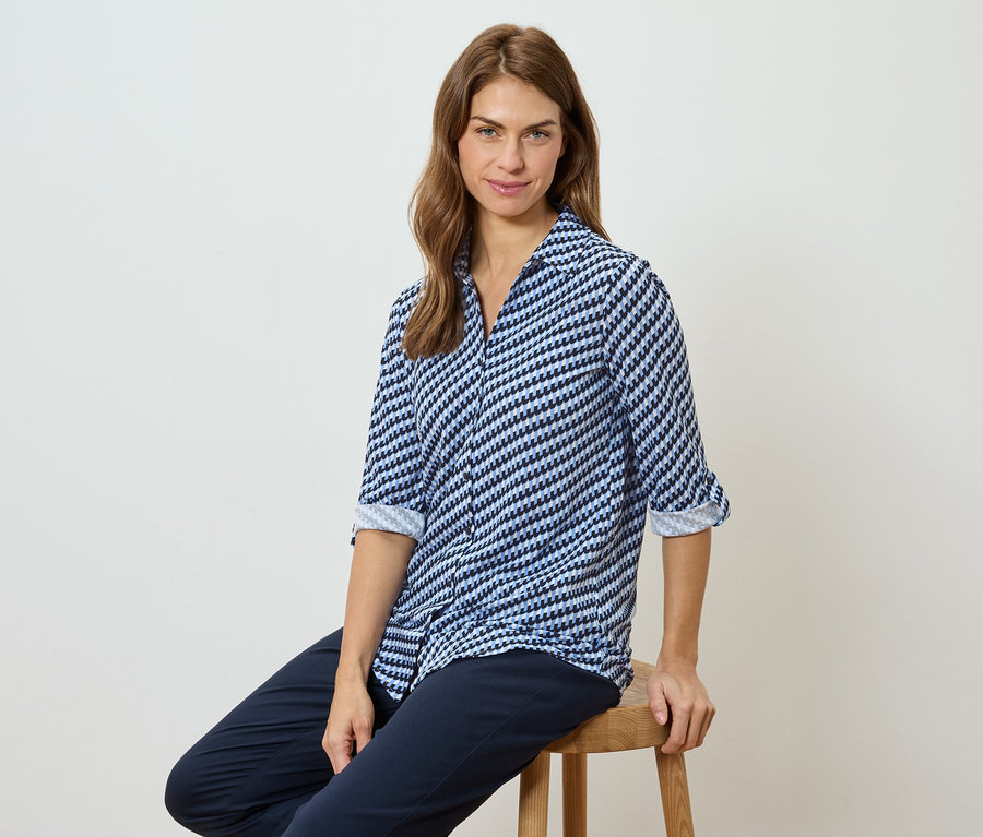 Kobieta siedzi na stołku, ubrana w bluzkę w niebiesko-biały wzór i ciemne spodnie.
