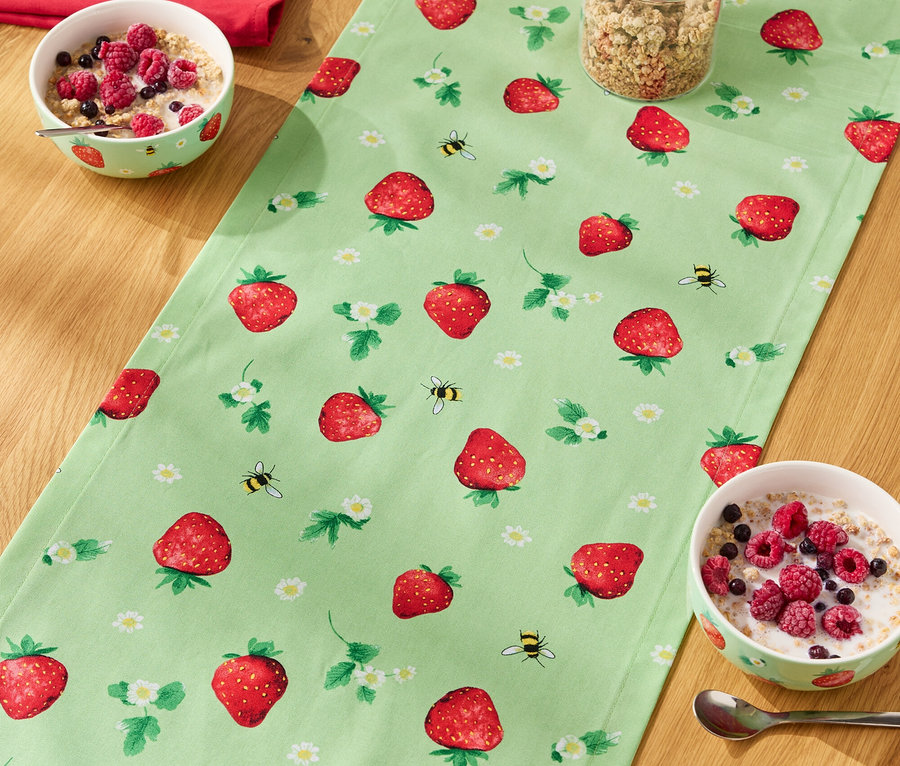 Na drewnianym stole leży bieżnik w truskawki, na nim miski z płatkami, szklanka musli i butelka mleka.