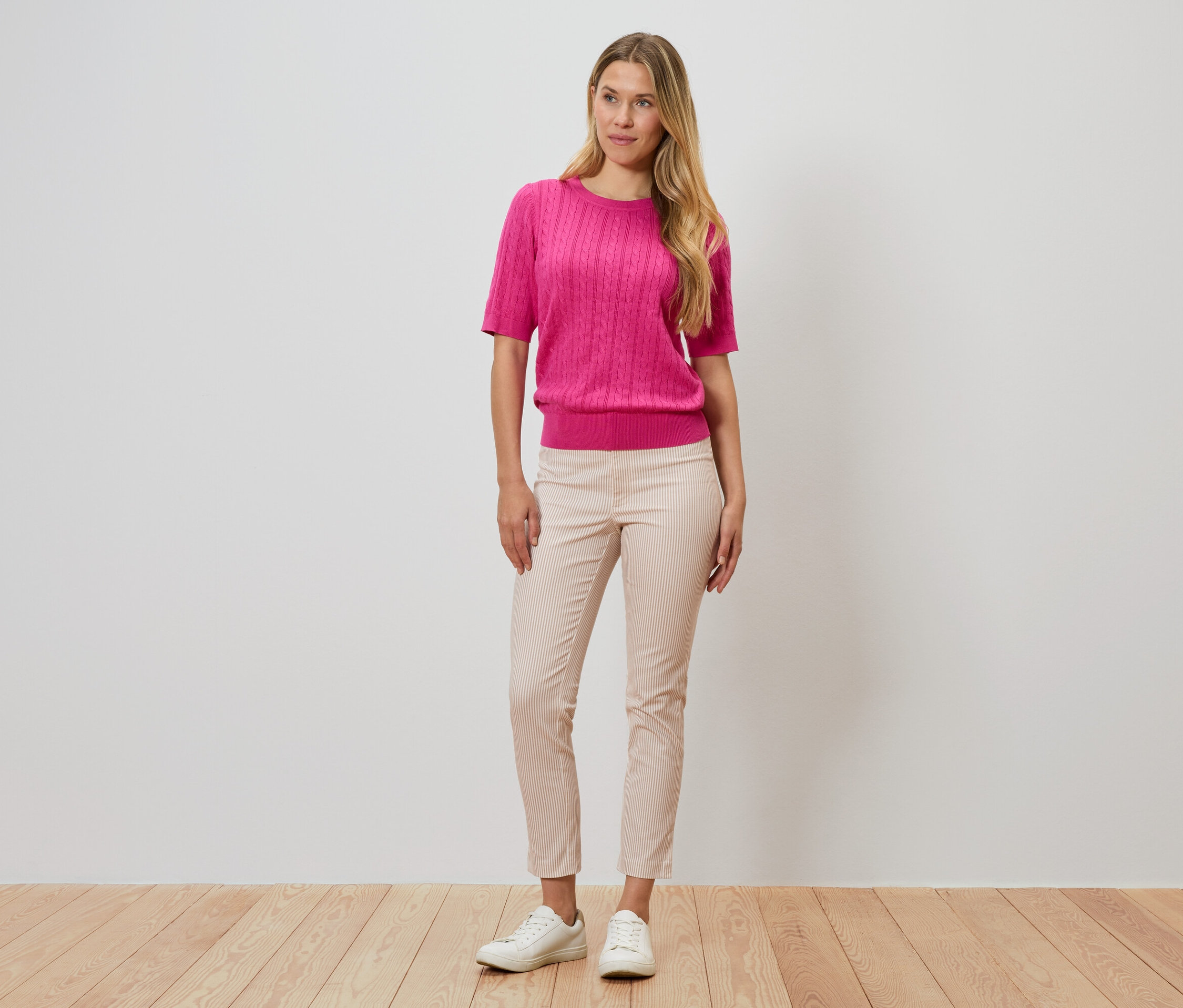 Kobieta stoi na tle ściany, ubrana w różowy sweter, prążkowane spodnie i białe buty.