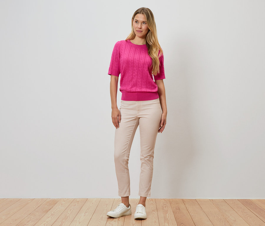 Kobieta stoi na tle ściany, ubrana w różowy sweter, prążkowane spodnie i białe buty.