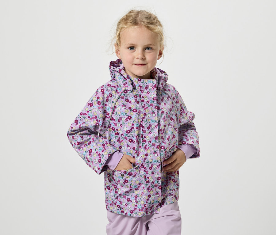 Dziewczynka z blond włosami w liliowej kurtce w kwiaty stoi na białym tle.