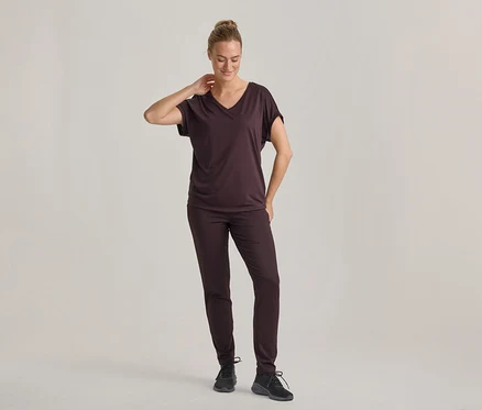 Kobieta w ciemnobrązowym T-shircie z dekoltem w serek i pasujących spodniach, w czarnych sneakersach, stoi i patrzy w dół, z ręką na karku.