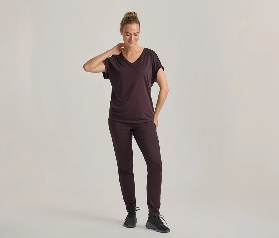 Kobieta w ciemnobrązowym T-shircie z dekoltem w serek i pasujących spodniach, w czarnych sneakersach, stoi i patrzy w dół, z ręką na karku.