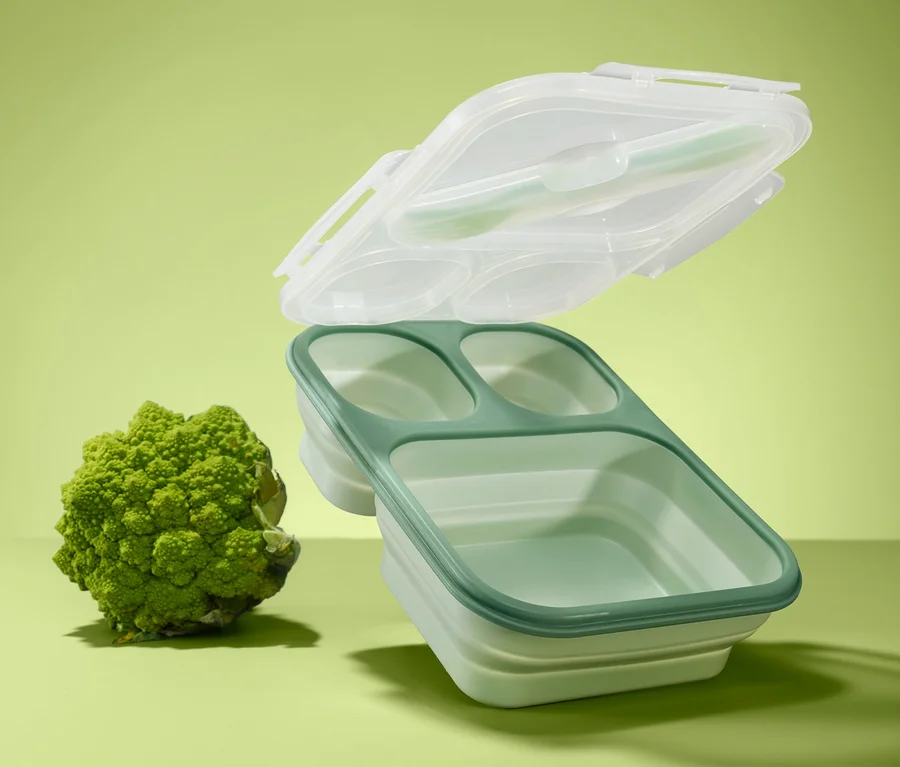 Zielony lunchbox składany z trzema przegródkami i przezroczystą pokrywką unosi się nad brokułem na zielonym tle.