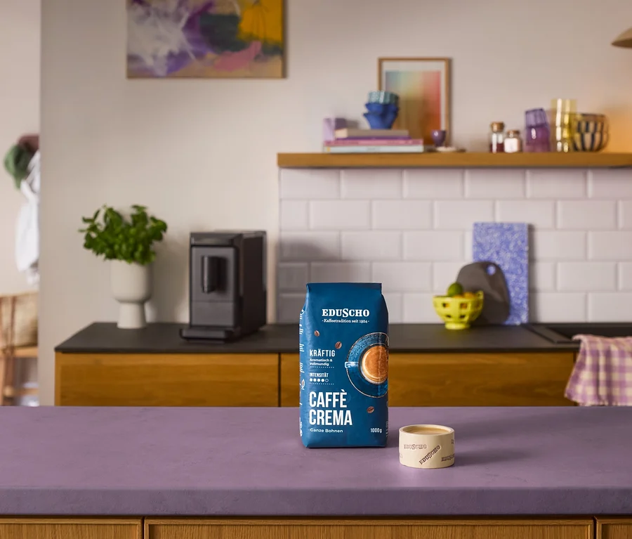 Niebieska paczka kawy Eduscho Caffè Crema i mała filiżanka Eduscho na fioletowym blacie przed czarnym automatycznym ekspresem do kawy »Esperto mini« w kuchni.