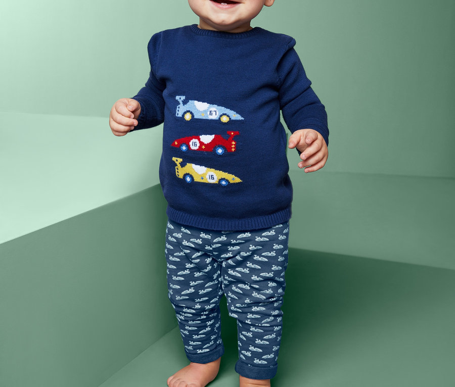 Uśmiechnięte dziecko stoi ubrane w niebieski sweter z samochodami i spodnie dresowe.