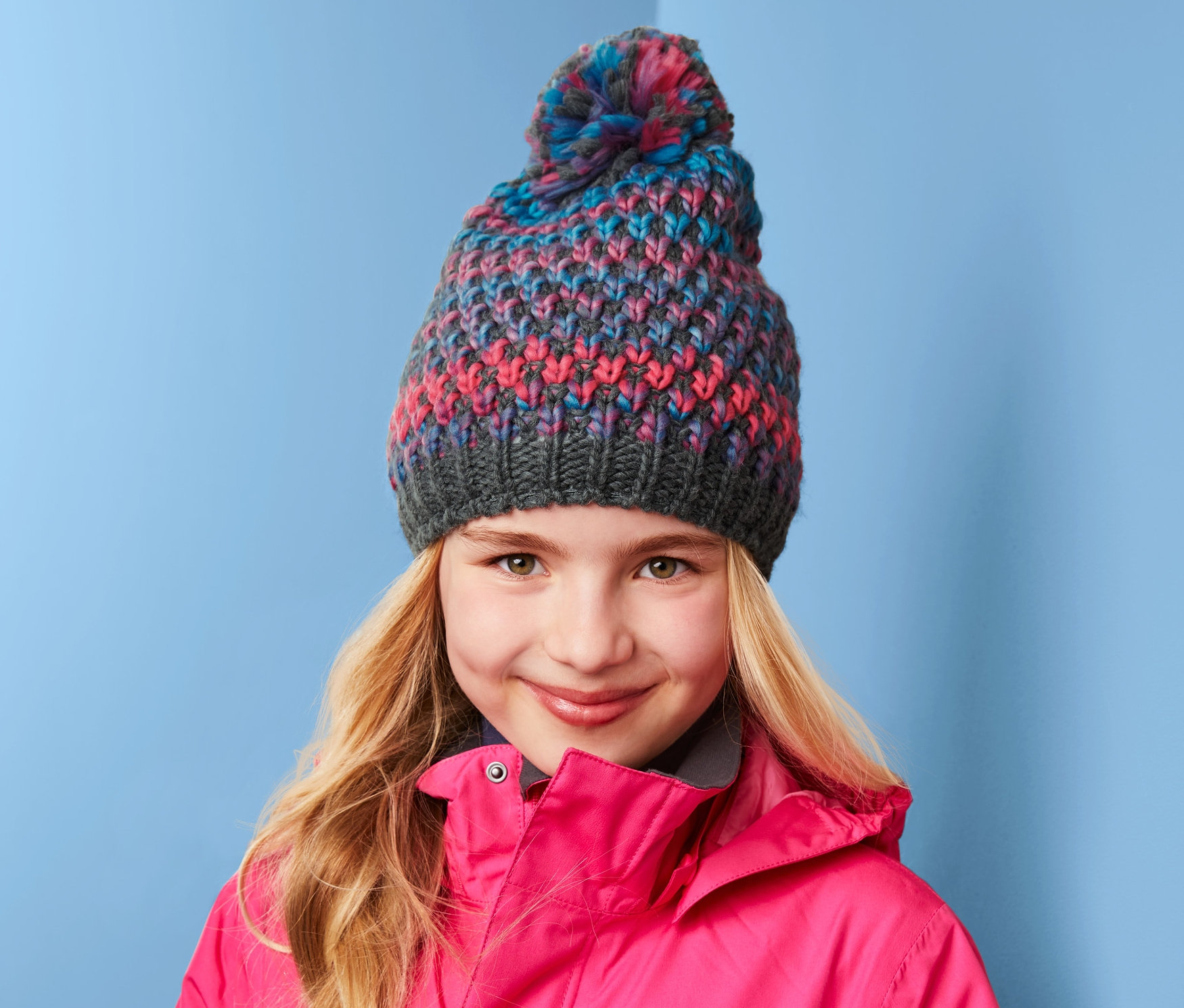 Dziewczyna z blond włosami w dziewczęcej czapce z dzianiny i różowej kurtce na tle niebieskiego tła.