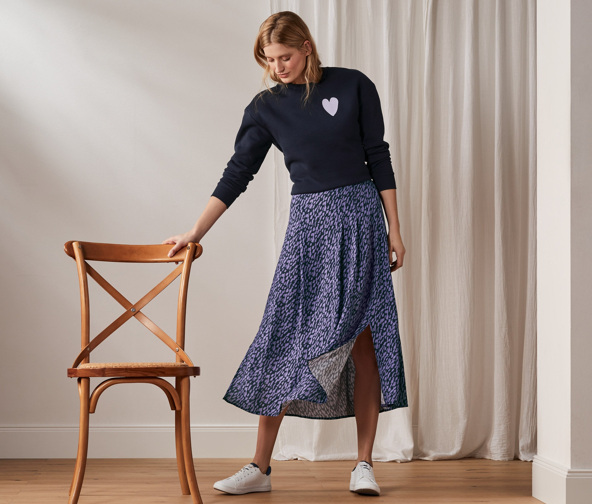 Kobieta stoi oparta o drewniane krzesło, ubrana w spódnicę z nadrukiem, granatowy sweter i białe tenisówki.