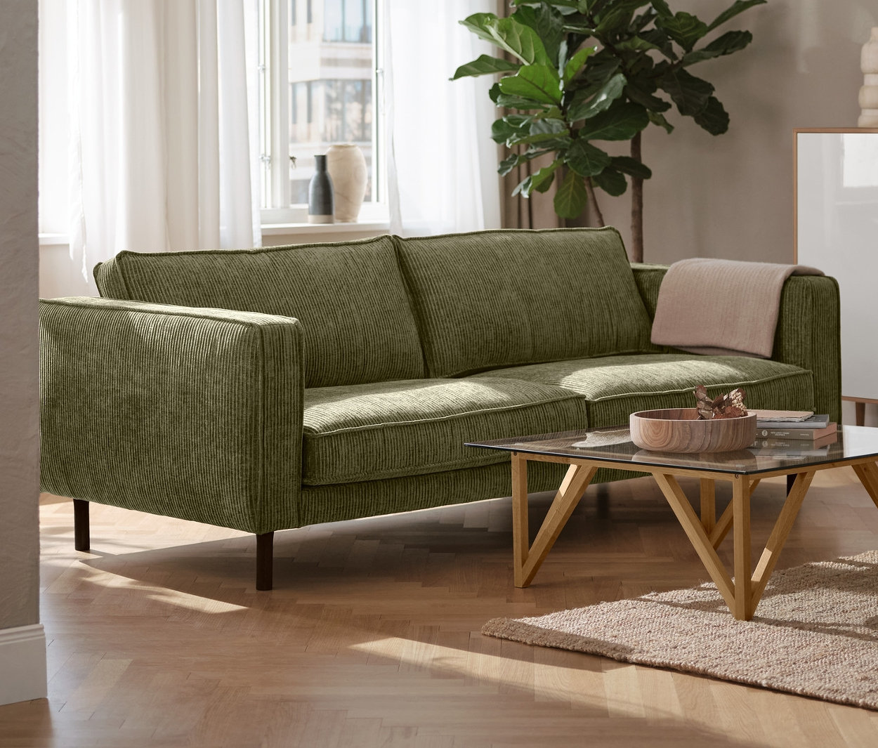 Zielona 3-osobowa sofa sztruksowa w salonie, obok czarna lampa sufitowa z metalu i biała szafka typu highboard »Bekeson«.
