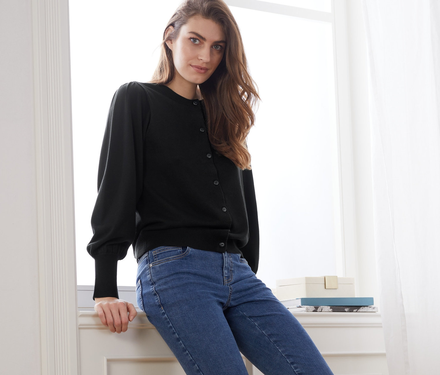 Kobieta siedzi na parapecie w czarnym swetrze z delikatnej dzianiny z bufiastymi rękawami i niebieskich dżinsach.