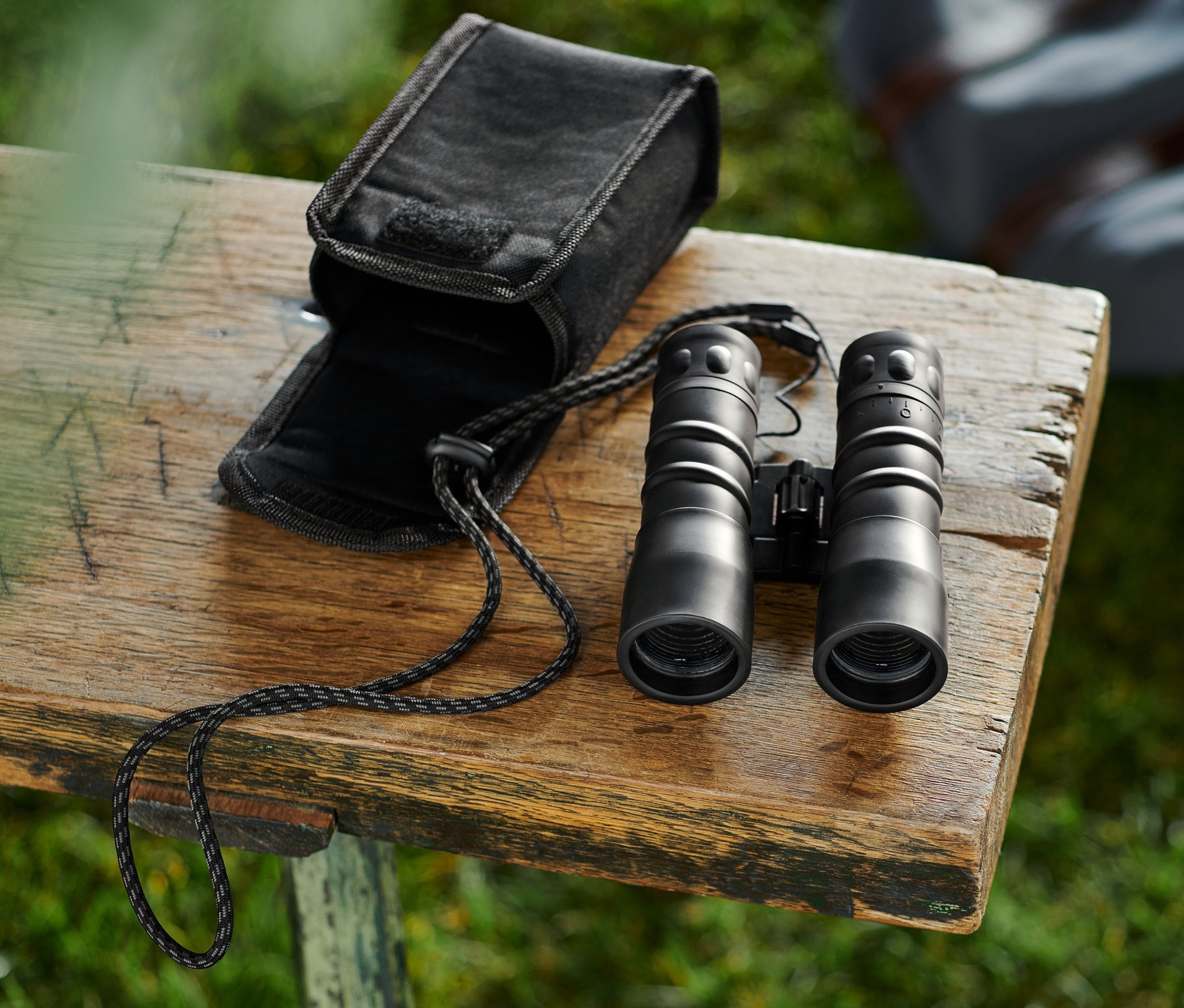 Czarne lornetki kieszonkowe »12x32« leżą obok otwartego futerału na drewnianej ławce.