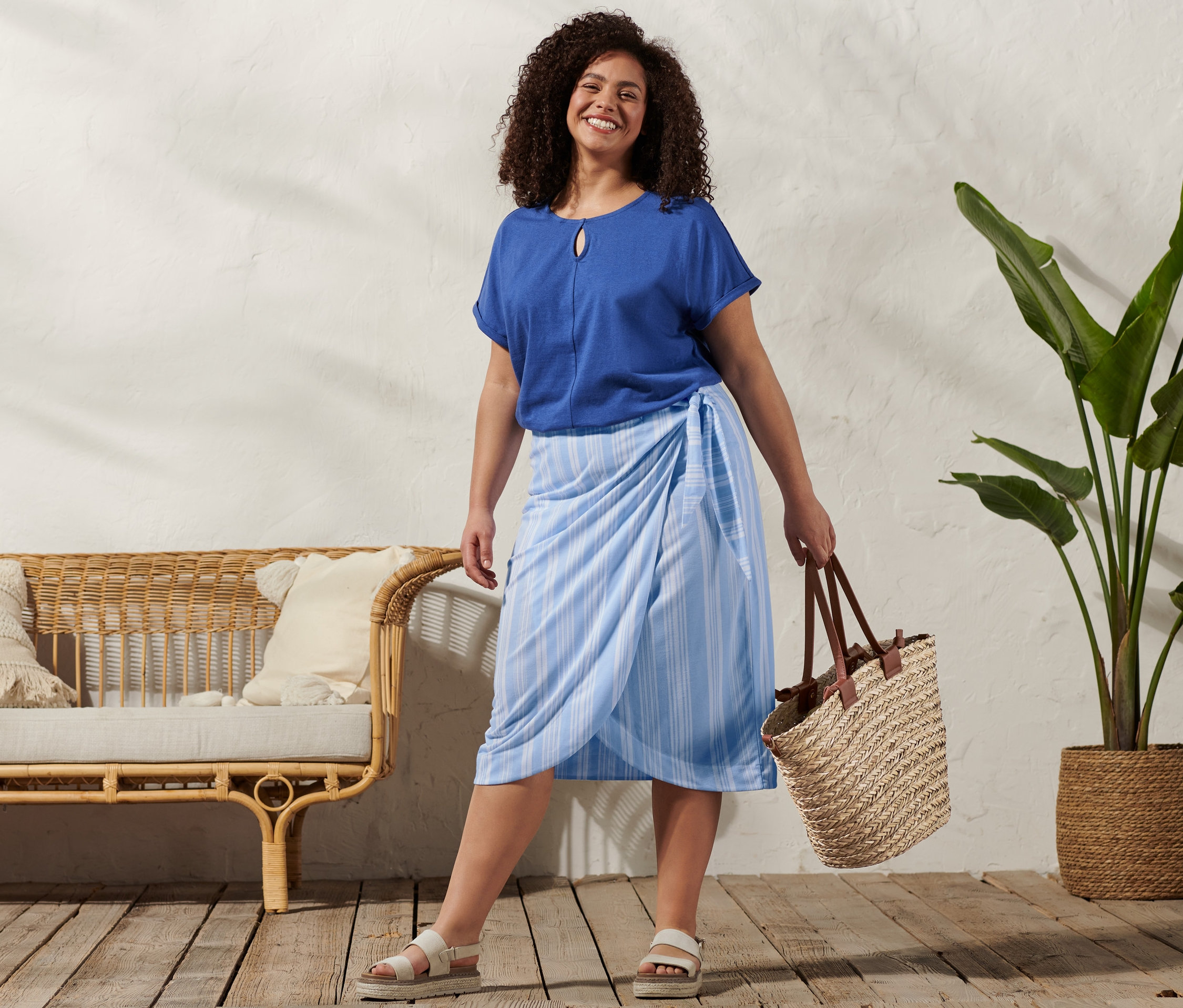 Kobieta ma na sobie bluzkę z mieszanki bawełny i lnu, spódnicę kopertową w paski, sandały skórzane na platformie i torbę z plecionki.