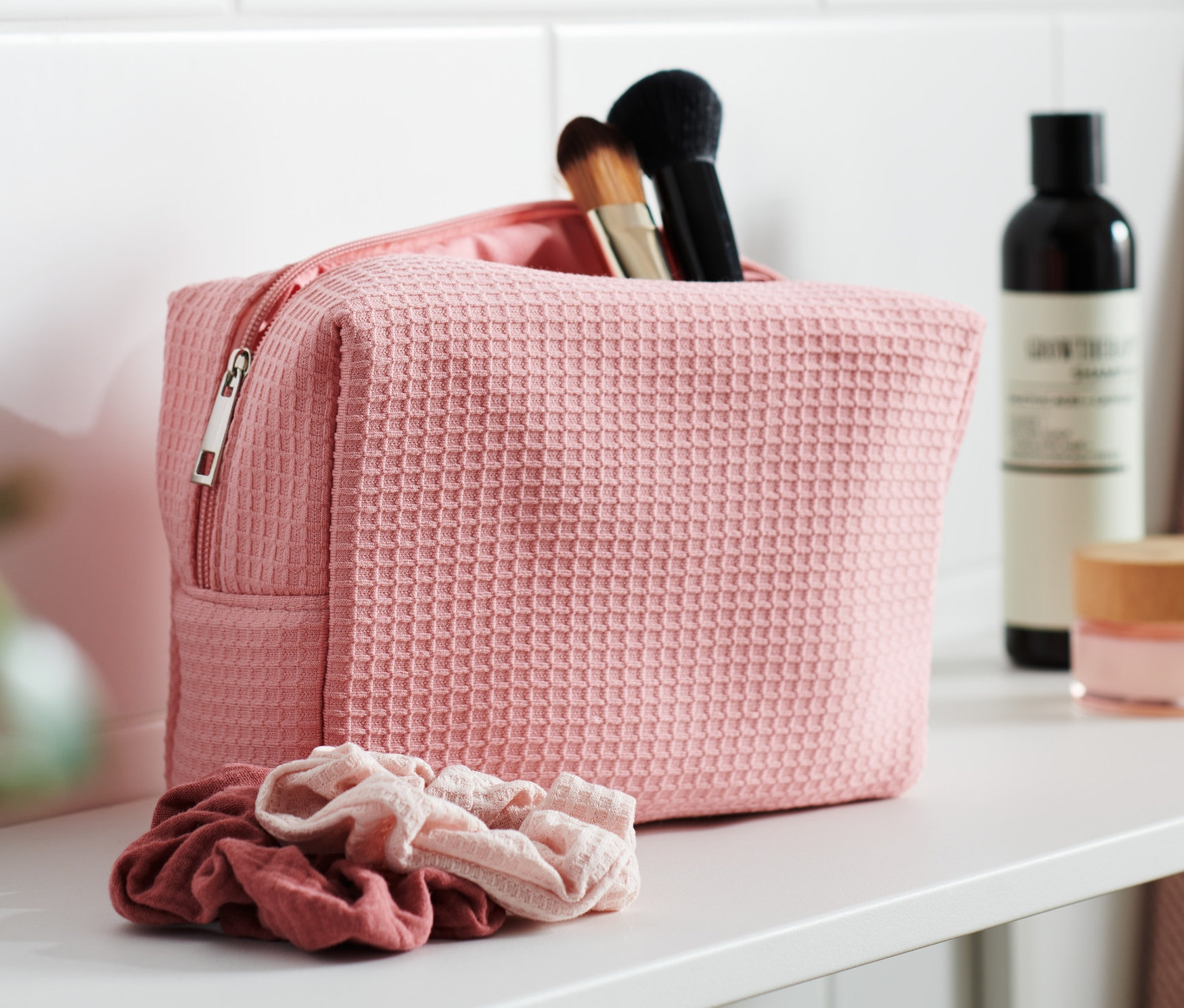 Różowa kosmetyczka z pędzlami do makijażu na białej półce. Obok niej leżą dwie gumki do włosów.