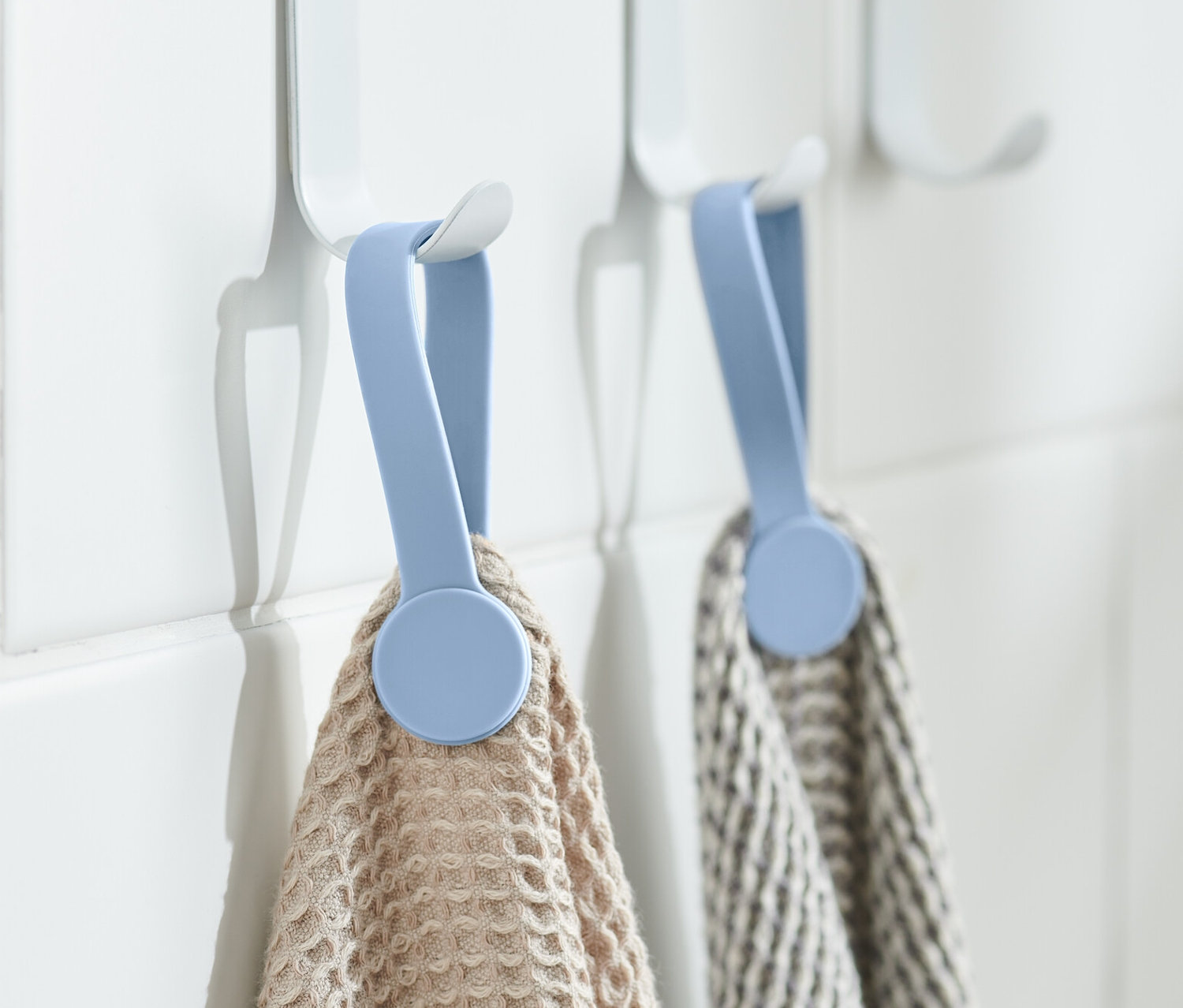 Dwa ręczniki wiszą na białych haczykach z niebieskimi klipsami magnetycznymi, 2 sztuki, na białej ścianie.