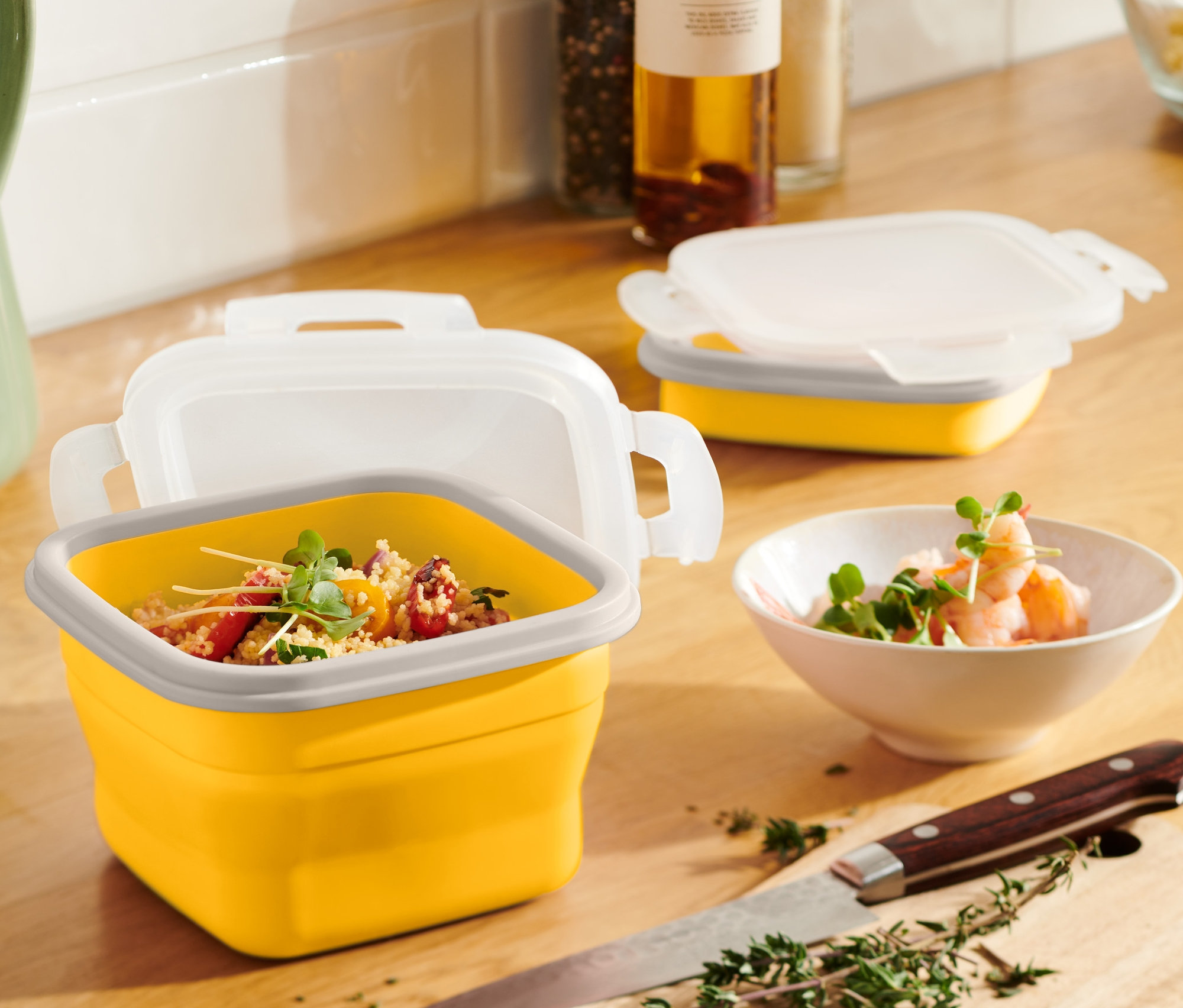 Żółty składany pojemnik na żywność z zawartością stoi na drewnianym stole obok zielonego dzbanka i miski z krewetkami.