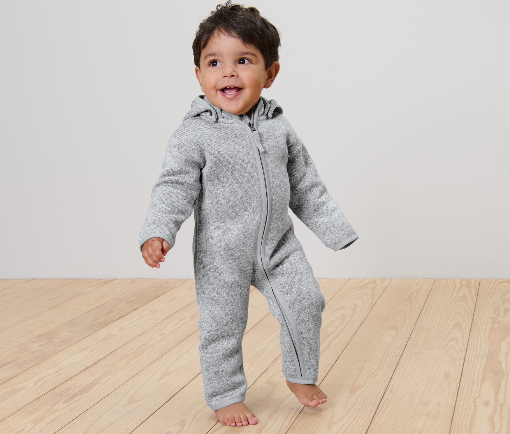Uśmiechnięte dziecko stoi na drewnianej podłodze, ubrane w szary dzianinowy kombinezon.