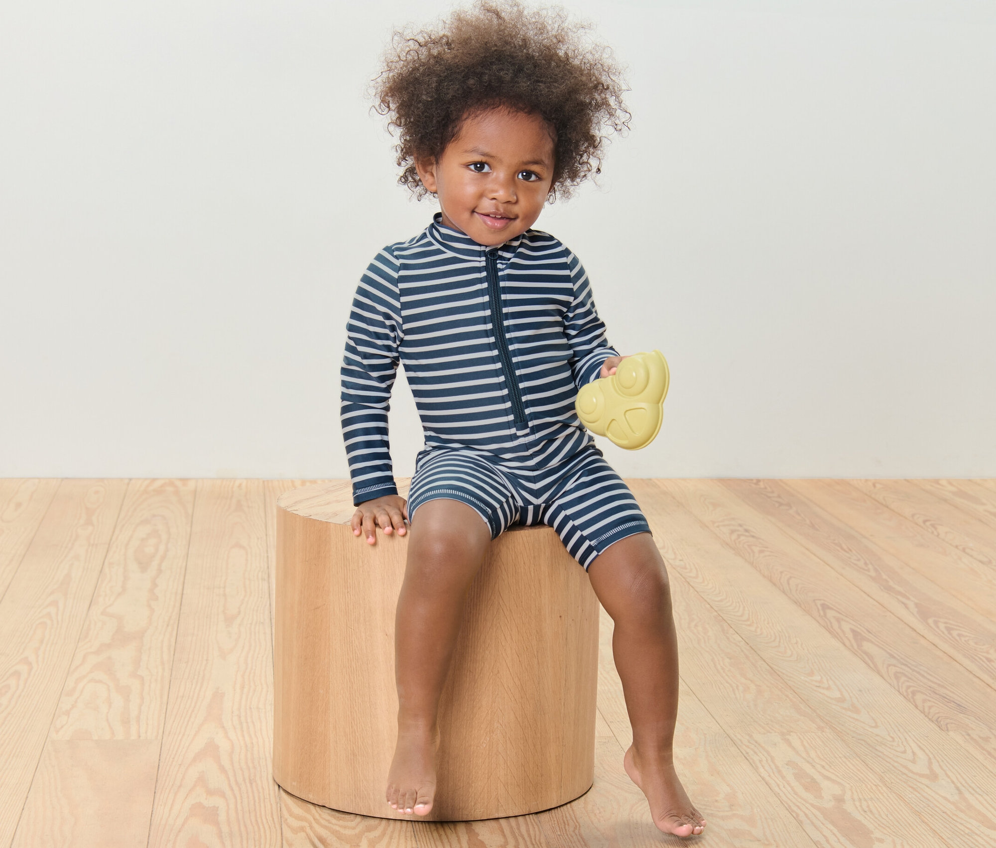 Dziecko siedzi na drewnianym stołku i trzyma żółtą zabawkę, ubrane jest w kombinezon niemowlęcy z filtrem UV 80, w paski.