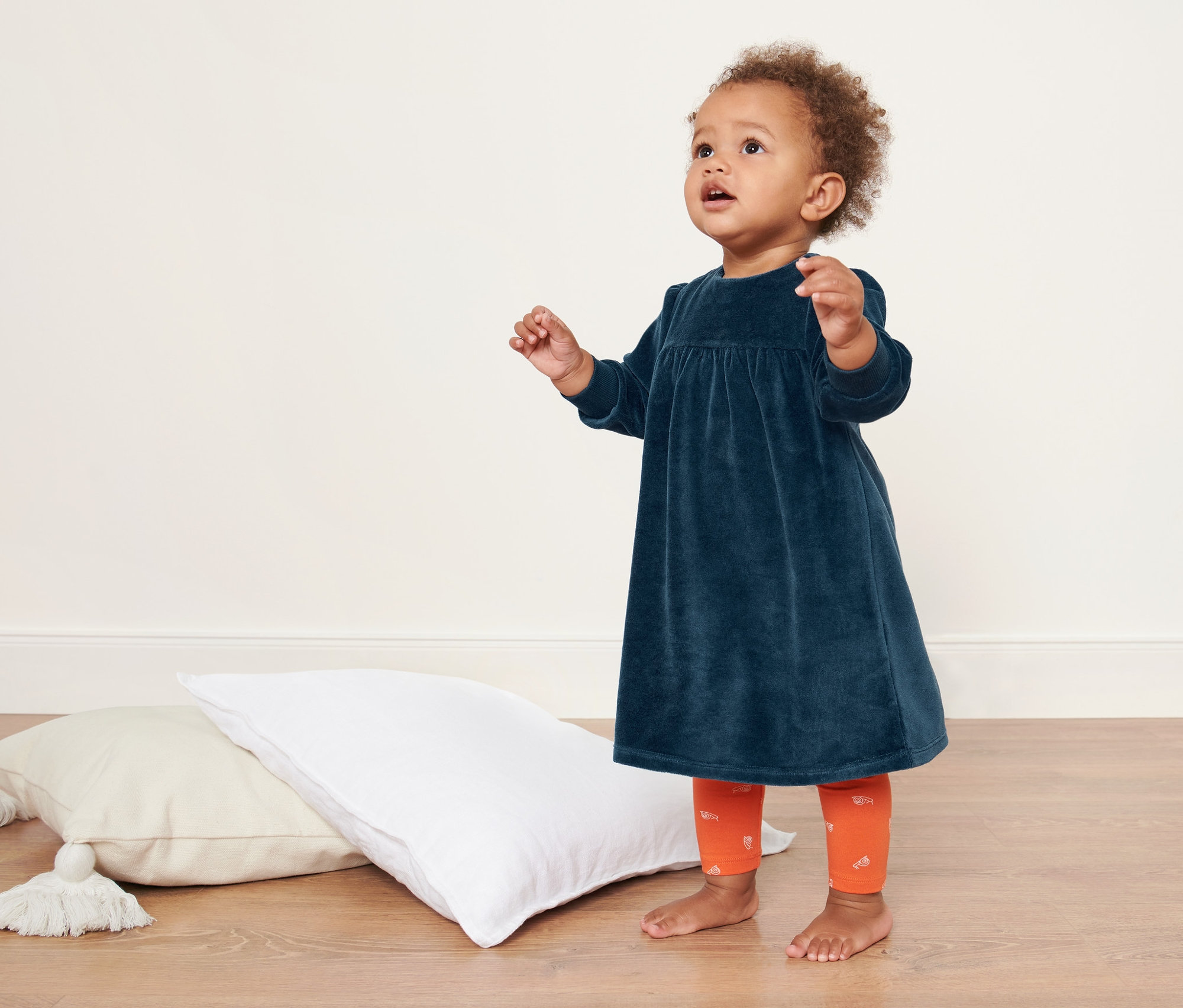 Dziecko stoi na podłodze obok poduszek, ubrane w sukienkę welurową.
