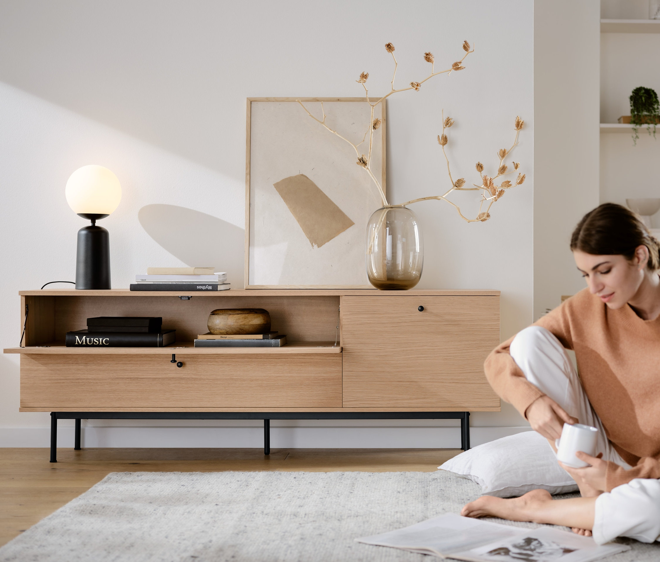 Kobieta siedzi na podłodze obok szafki pod telewizor »Finnsbo« i lampy stołowej ze szklaną kulą, czarna.