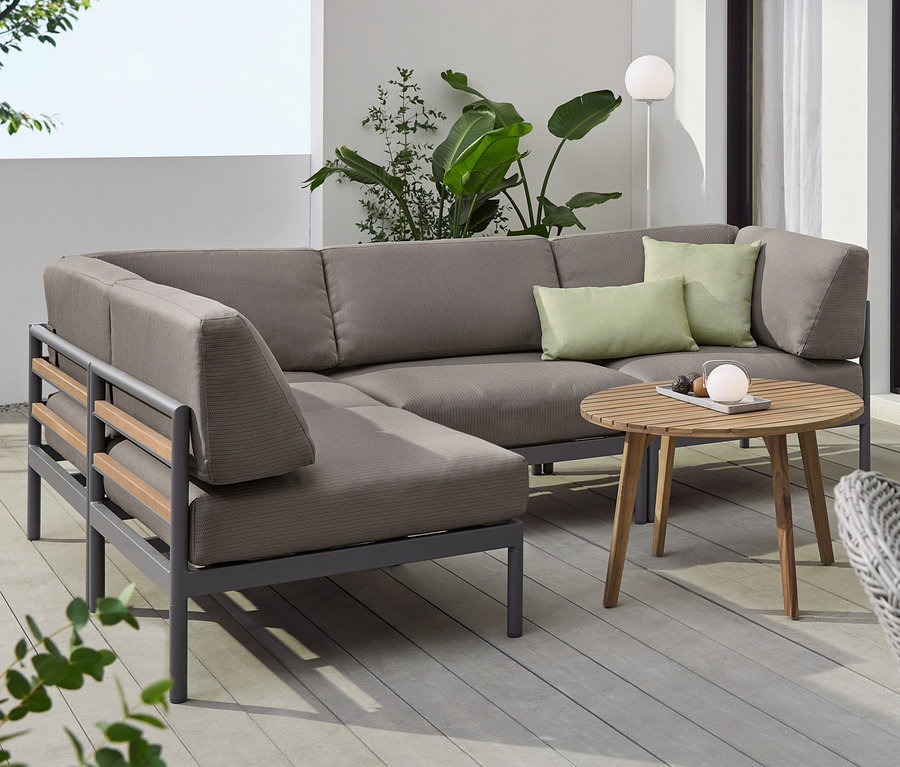 Szara sofa narożna z tapicerką premium, okrągły stolik kawowy i stolik pomocniczy „Lenja” z 2 poziomami. Na stołach stoją akumulatorowe lampy LED.
