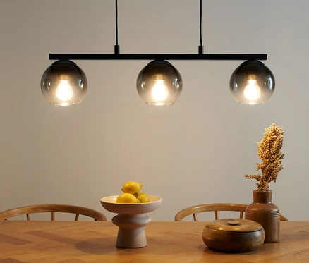Lampa wisząca z kloszami z przydymionego szkła nad drewnianym stołem. Na stole misa z owocami i drewniana puszka.