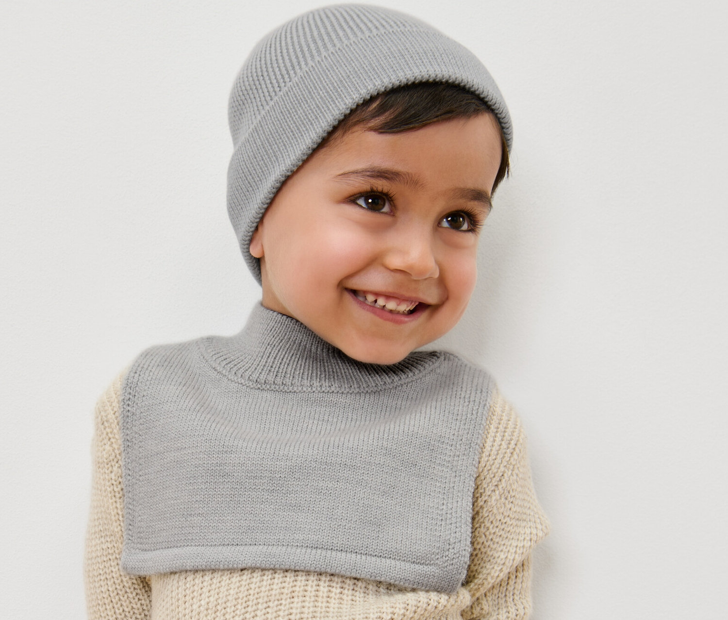 Uśmiechnięty chłopiec ma na sobie szary kołnierz z golfem z wełny i czapkę.