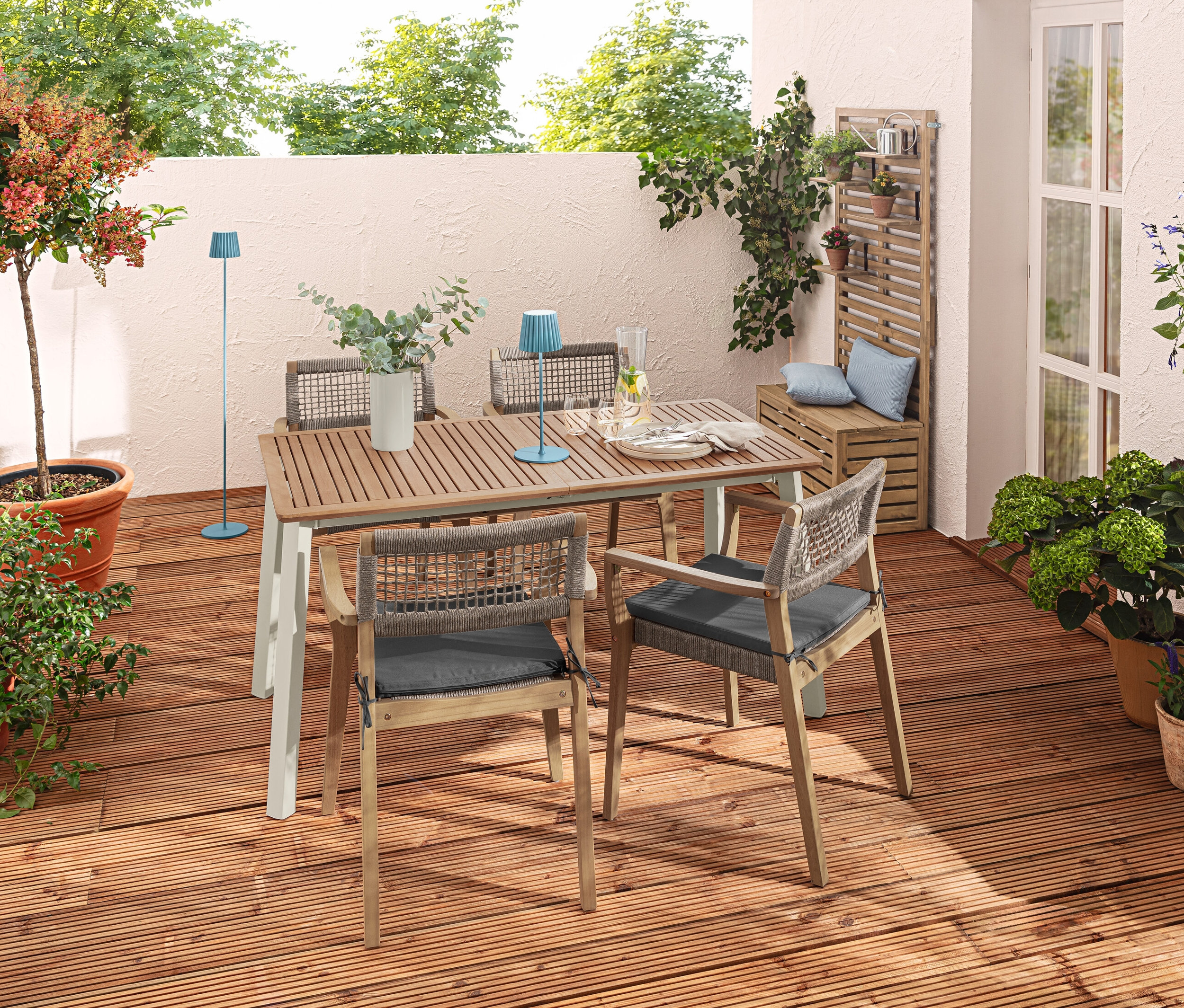 Zewnętrzny taras z rozsuwanym stołem „Liska”, krzesłami z możliwością układania jeden na drugim, niebieską akumulatorową lampą stołową i poduszkami ogrodowymi.