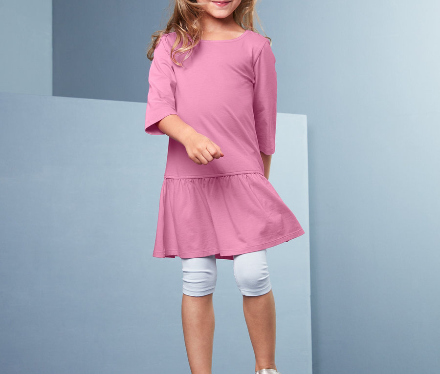 Dziewczynka stoi na szarym sześcianie i ma na sobie różową sukienkę dżersejową.