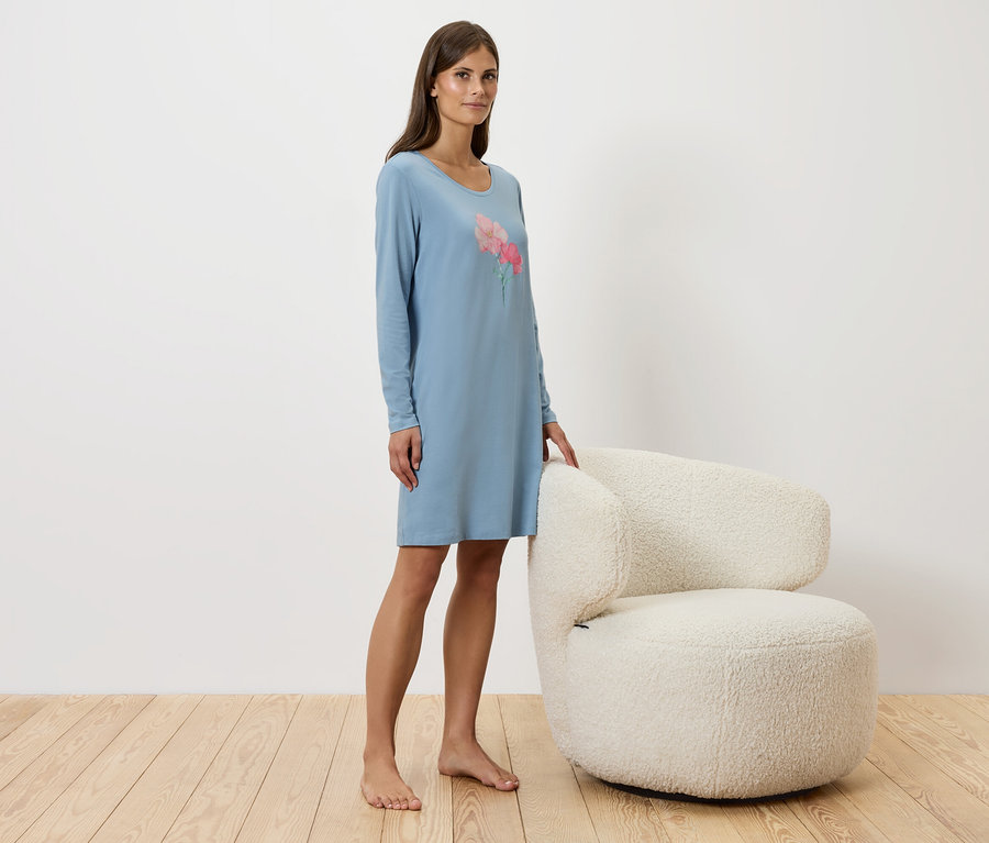 Kobieta stoi obok białego krzesła, ubrana w niebieską koszulę nocną.