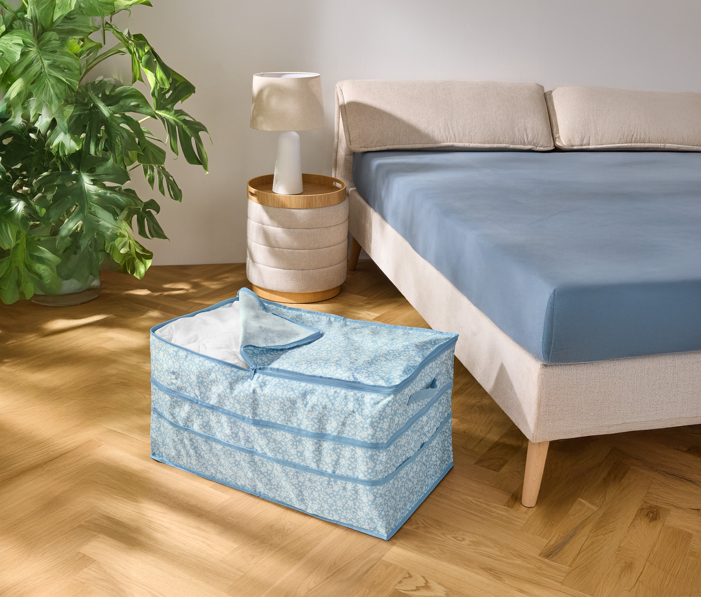 Na drewnianej podłodze stoi jasnoniebieski wzorzysty pojemnik do przechowywania obok łóżka z niebieskim prześcieradłem.