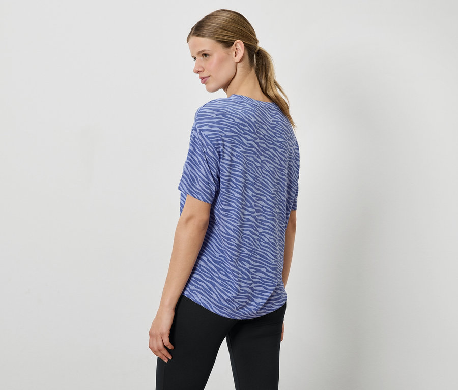 Kobieta w niebieskim topie we wzór zwierzęcy i czarnych spodniach, patrzy przez ramię.