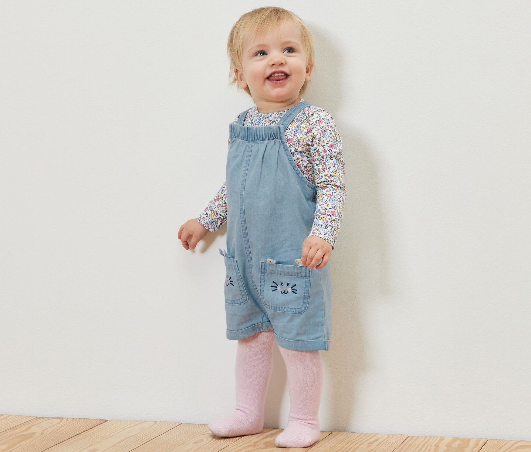 Małe dziecko stoi na drewnianej podłodze, ubrane w ogrodniczki z tkaniny chambray i koszulkę w kwiaty.