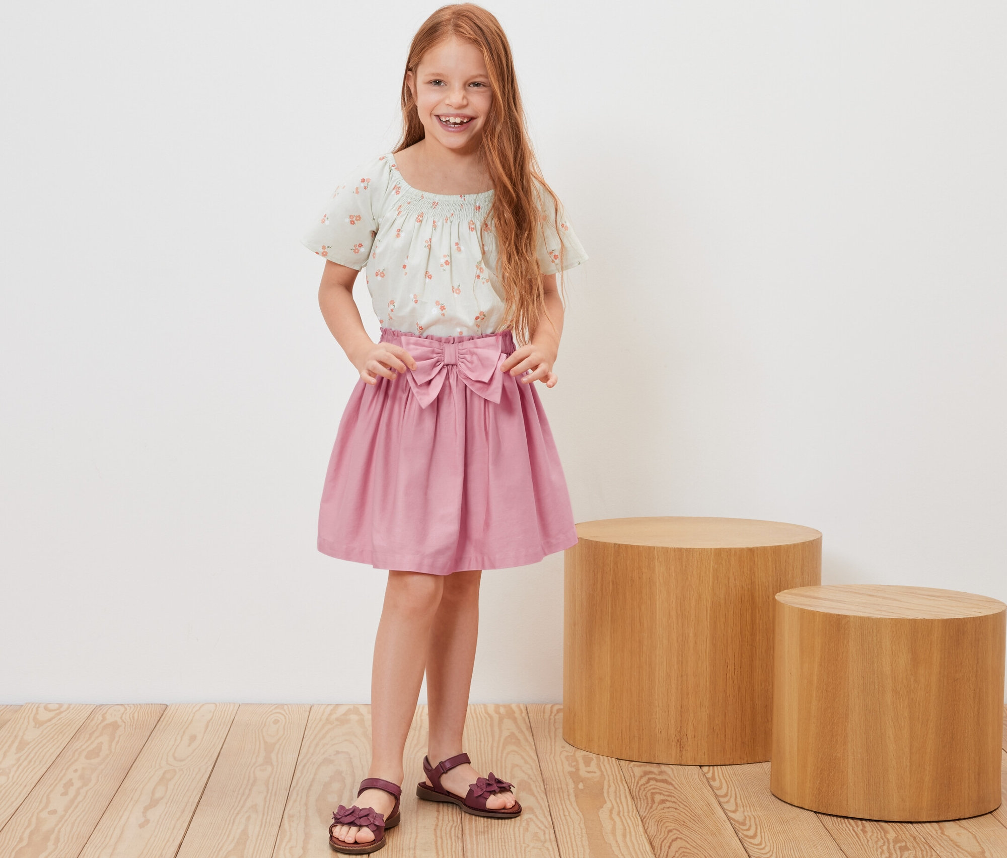 Uśmiechnięta, rudowłosa dziewczynka pozuje w spódnicy z tkaniny i kwiecistym topie.