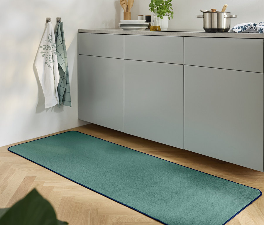 Zielony dywan leży na drewnianej podłodze w kuchni z szarą szafką.