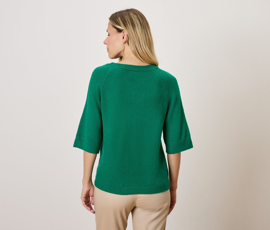 Kobieta ma na sobie zielony sweter z rękawami trzy czwarte i beżowe spodnie, stojąc tyłem do kamery.