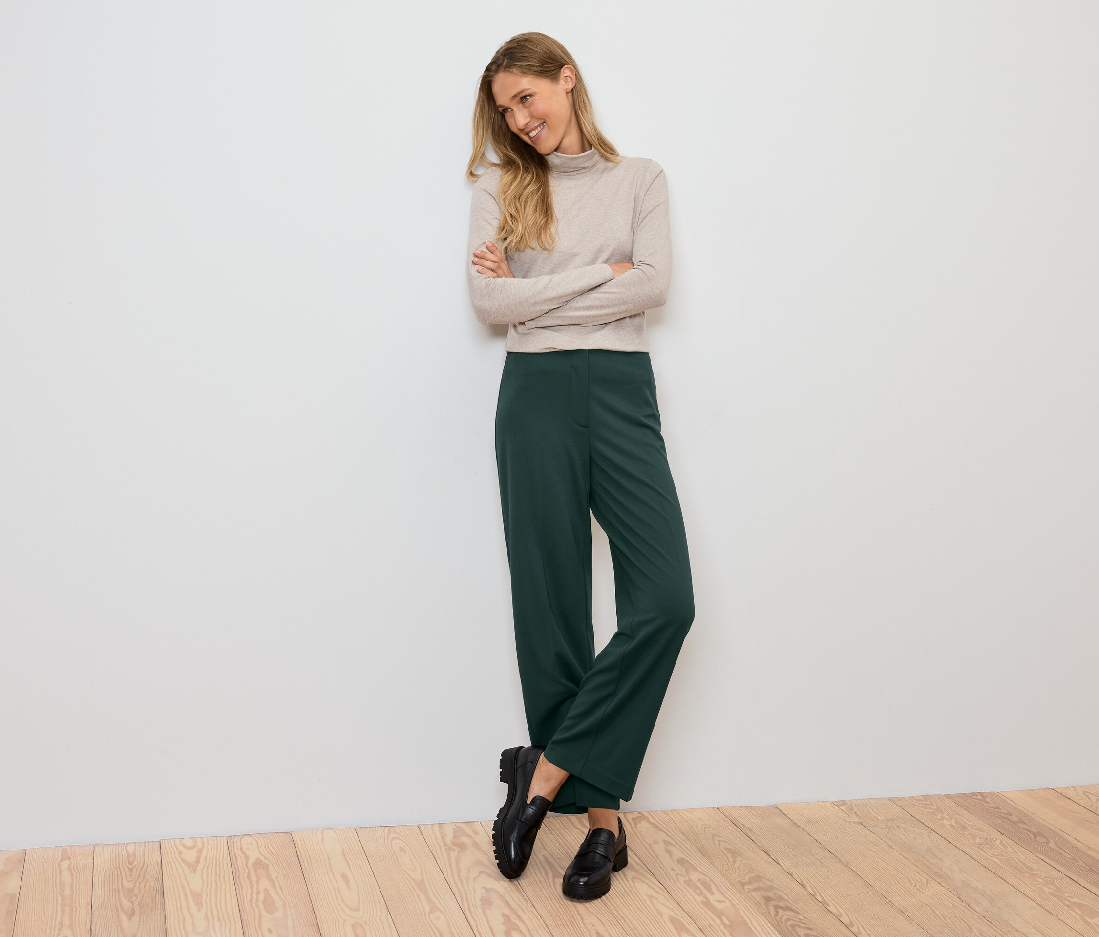 Uśmiechnięta kobieta pozuje z założonymi rękami na piersi, ubrana w beżowy golf, zielone spodnie i czarne buty.