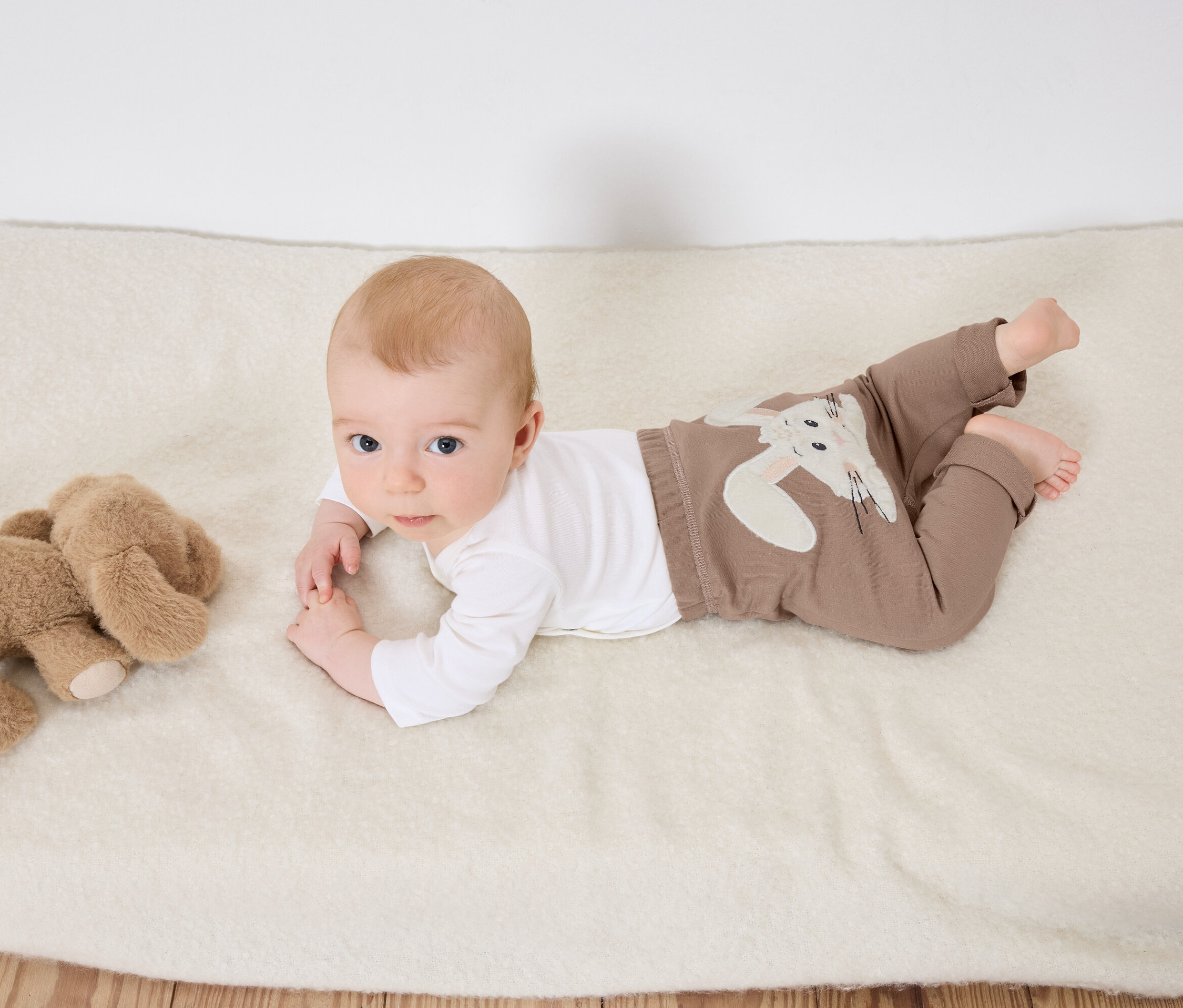 Niemowlę leży na brzuchu na białym kocu obok pluszowego słonia.
