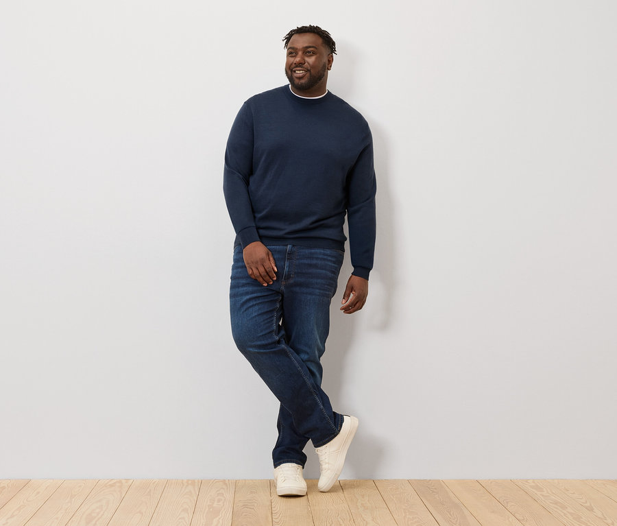 Mężczyzna pozuje oparty o ścianę, ubrany w granatowy sweter z wełny merino.