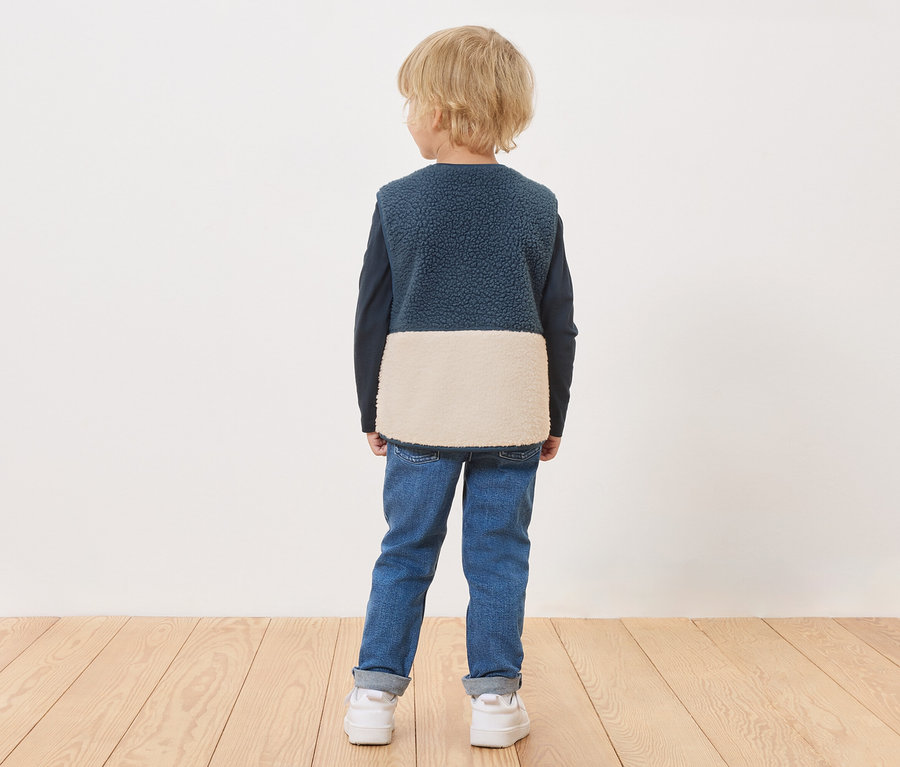 Chłopiec stoi tyłem, ma na sobie niebieską kamizelkę z kremowym dołem, niebieskie dżinsy i białe trampki.