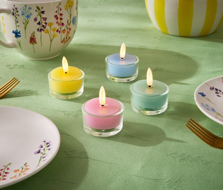 Na zielonym obrusie stoją cztery małe świeczki w pastelowych kolorach: żółtym, niebieskim, zielonym i różowym. Dodatkowo znajduje się tam talerz, filiżanka i miska z kwiatowym wzorem oraz dwa złote widelce.